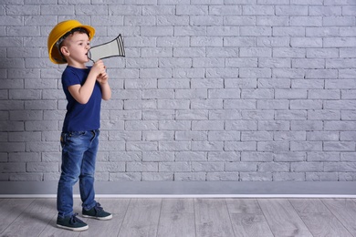 Adorable little boy in hardhat with paper megaphone on brick wall background