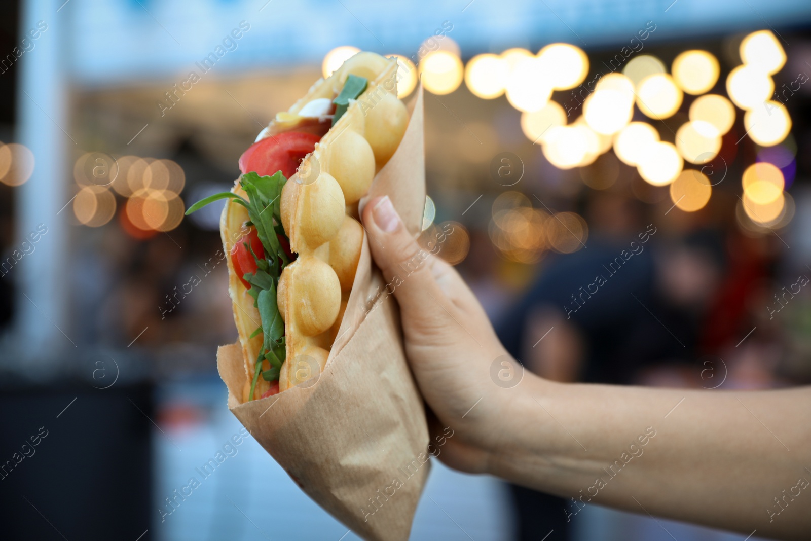 Photo of Young woman holding delicious bubble waffle with tomato and arugula outdoors, closeup