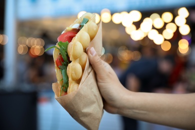 Photo of Young woman holding delicious bubble waffle with tomato and arugula outdoors, closeup