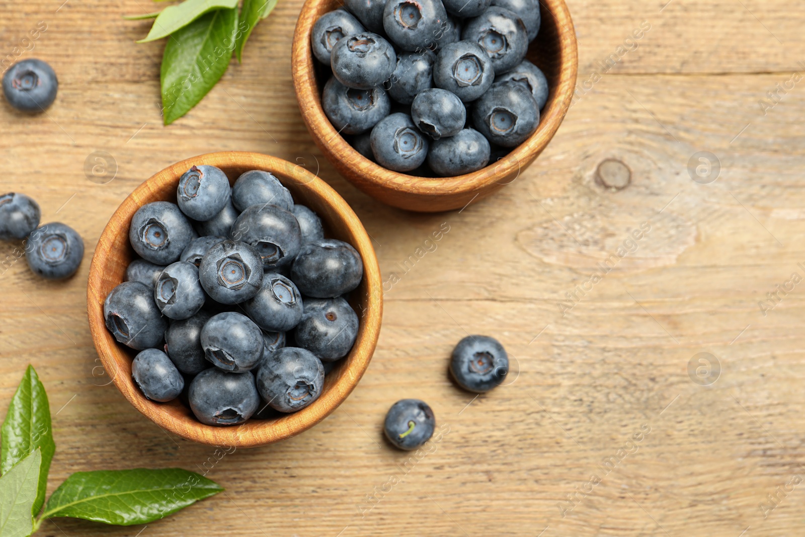 Photo of Bowls of fresh tasty blueberries and leaves on wooden table, flat lay. Space for text