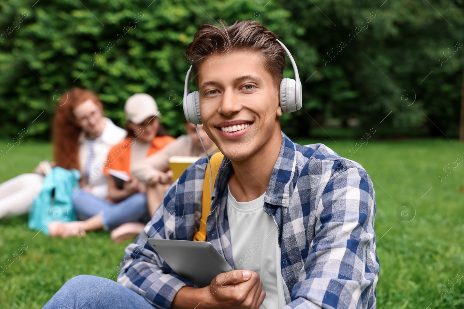 Photo of Students learning together in park. Happy young man with headphones working with tablet on green grass, selective focus