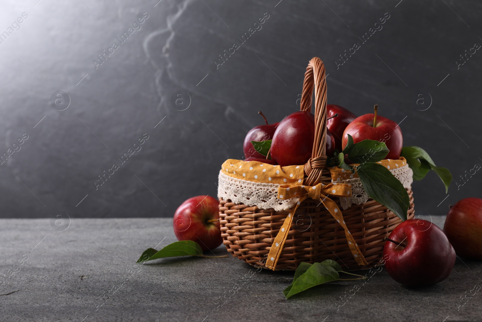 Photo of Ripe red apples and leaves in wicker basket on dark grey table. Space for text