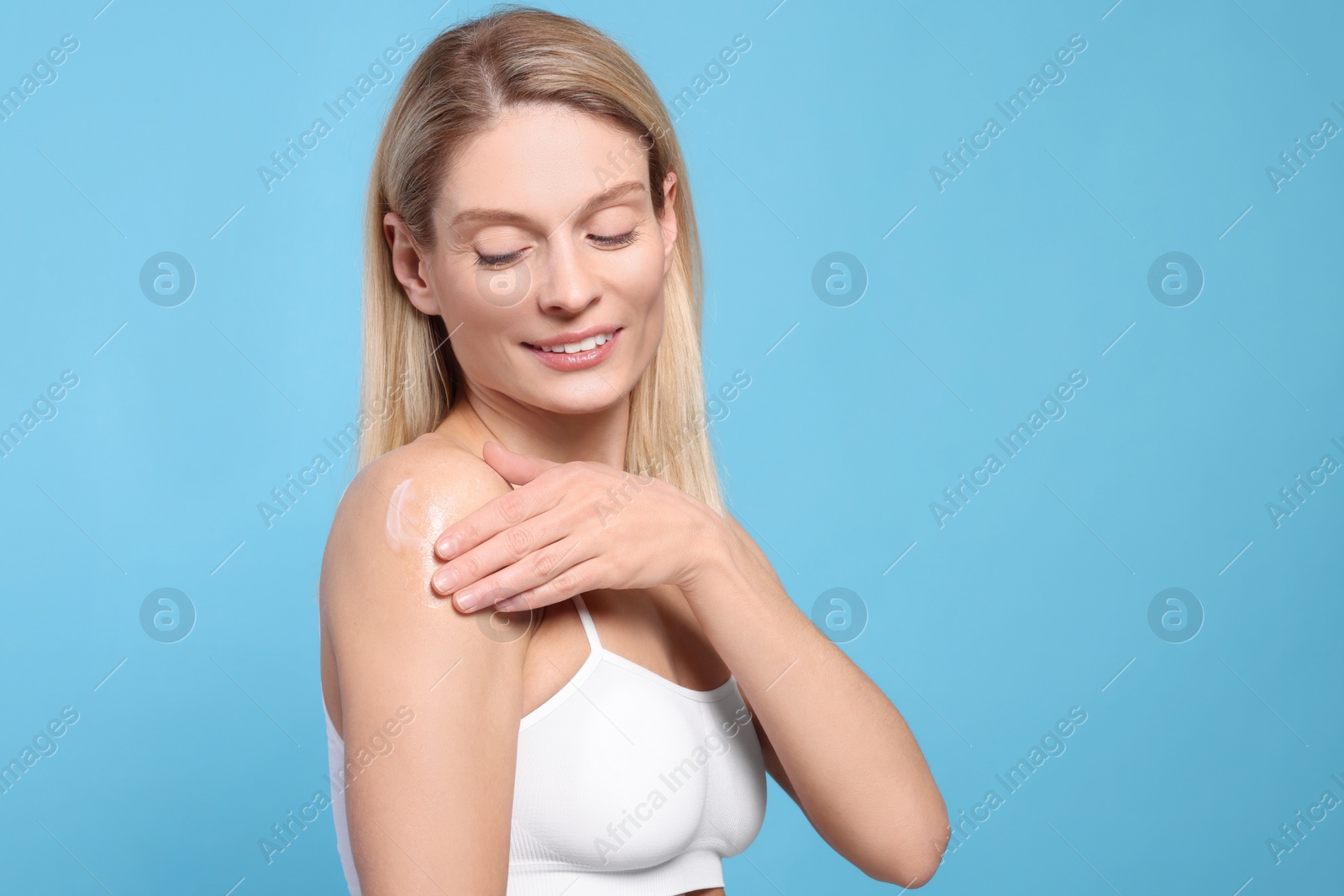 Photo of Woman applying body cream onto her arm against light blue background, space for text