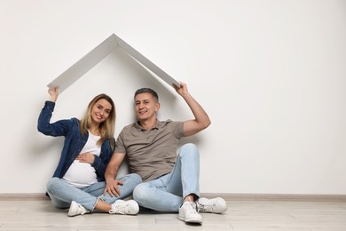 Photo of Young family housing concept. Pregnant woman with her husband sitting under cardboard roof on floor indoors. Space for text