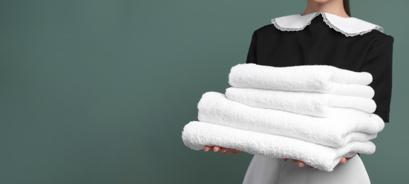 Young chambermaid holding stack of clean towels on color background, closeup view with space for text. Banner design