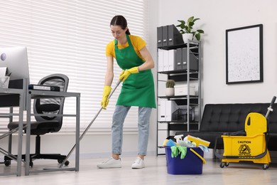 Photo of Cleaning service worker washing floor with mop. Bucket with supplies and wet floor sign in office