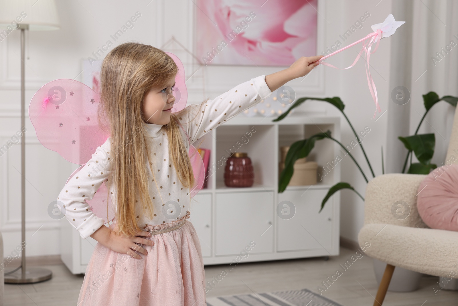 Photo of Cute little girl in fairy costume with pink wings and magic wand at home, space for text