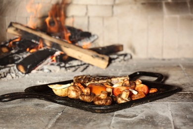 Photo of Grill pan with meat and garnish near burning firewood inside of oven
