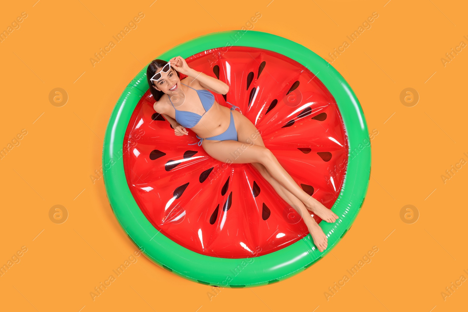 Photo of Happy young woman with beautiful suntan and sunglasses on inflatable mattress against orange background, top view