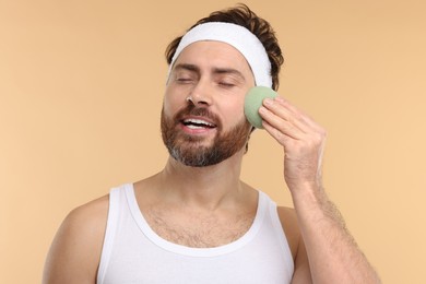 Photo of Man with headband washing his face using sponge on beige background