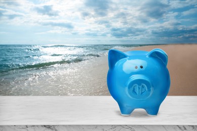 Saving money for summer vacation. Piggy bank on white marble surface near sandy beach and sea, space for text