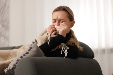 Photo of Sick woman blowing nose in tissue under blanket on sofa at home. Cold symptoms
