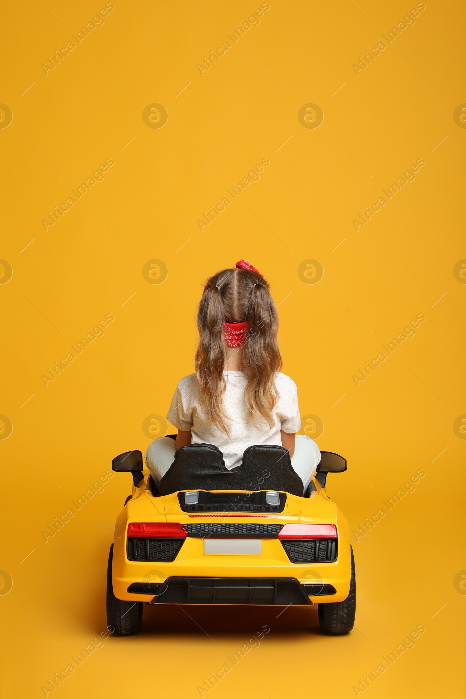Photo of Cute little girl driving children's electric toy car on yellow background, back view