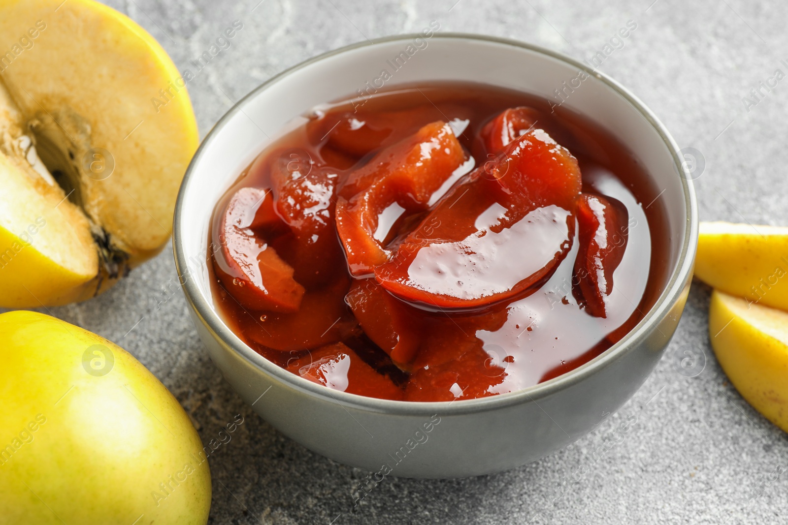 Photo of Tasty homemade quince jam in bowl and fruits on grey textured table, closeup