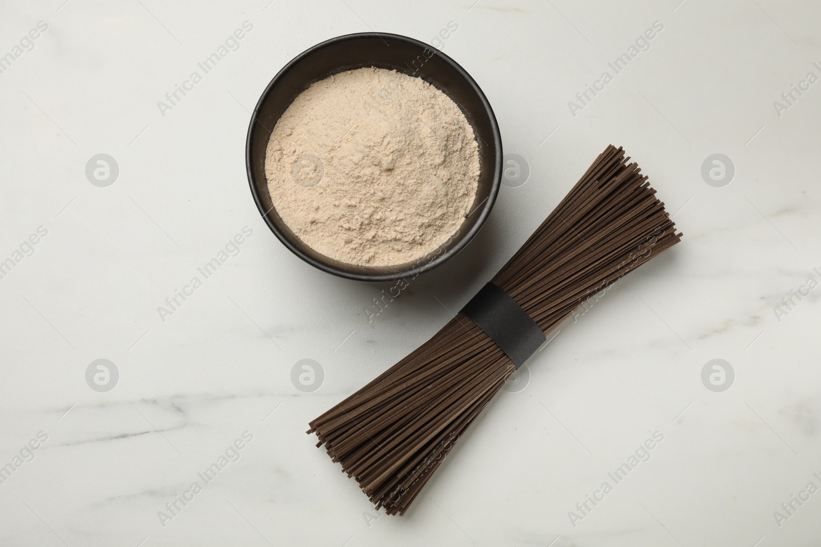 Photo of Uncooked buckwheat noodles (soba,) and flour on white marble table, top view