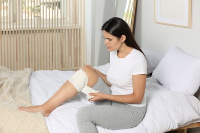 Young woman applying medical bandage onto knee in bedroom