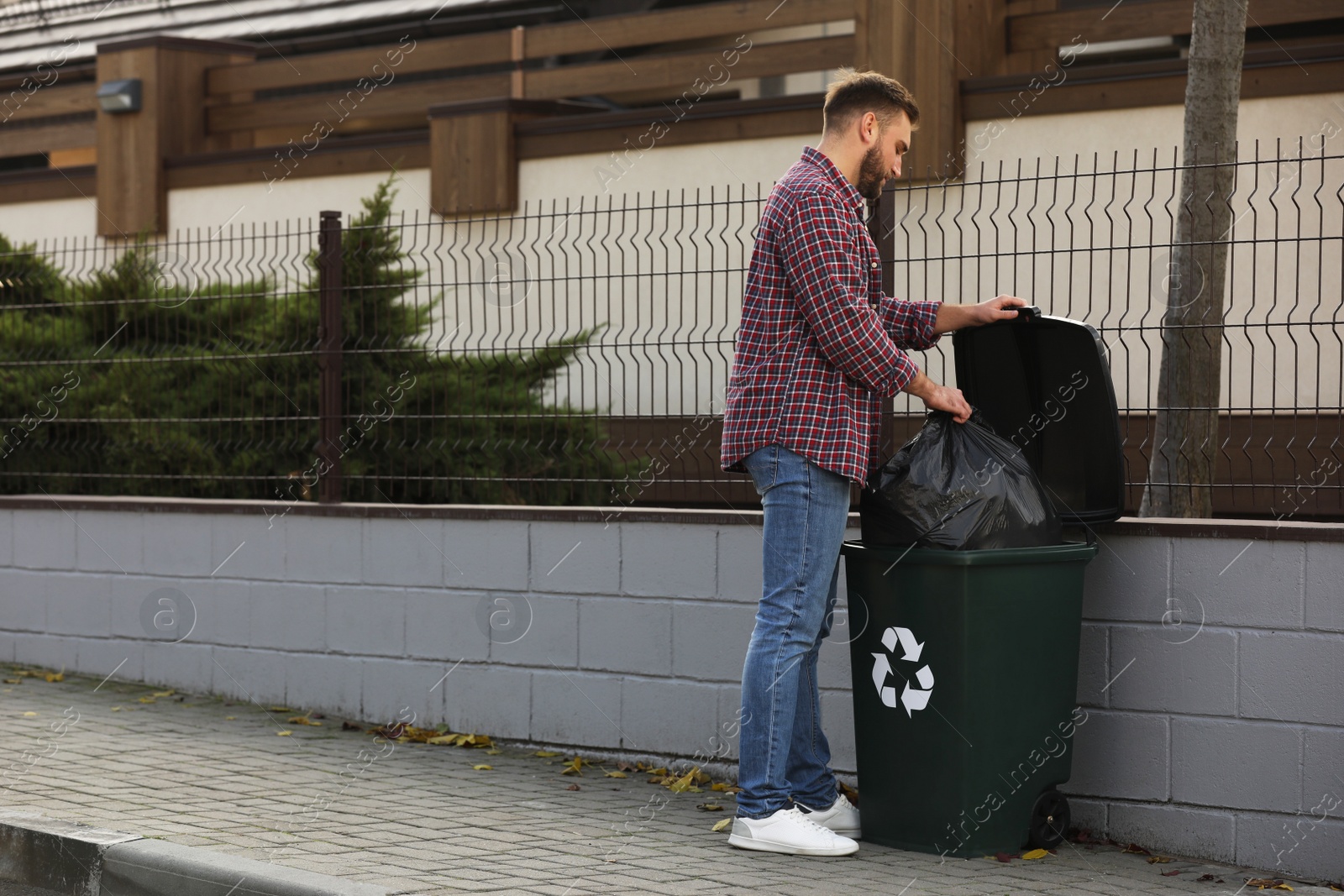 Photo of Man putting garbage bag into recycling bin outdoors