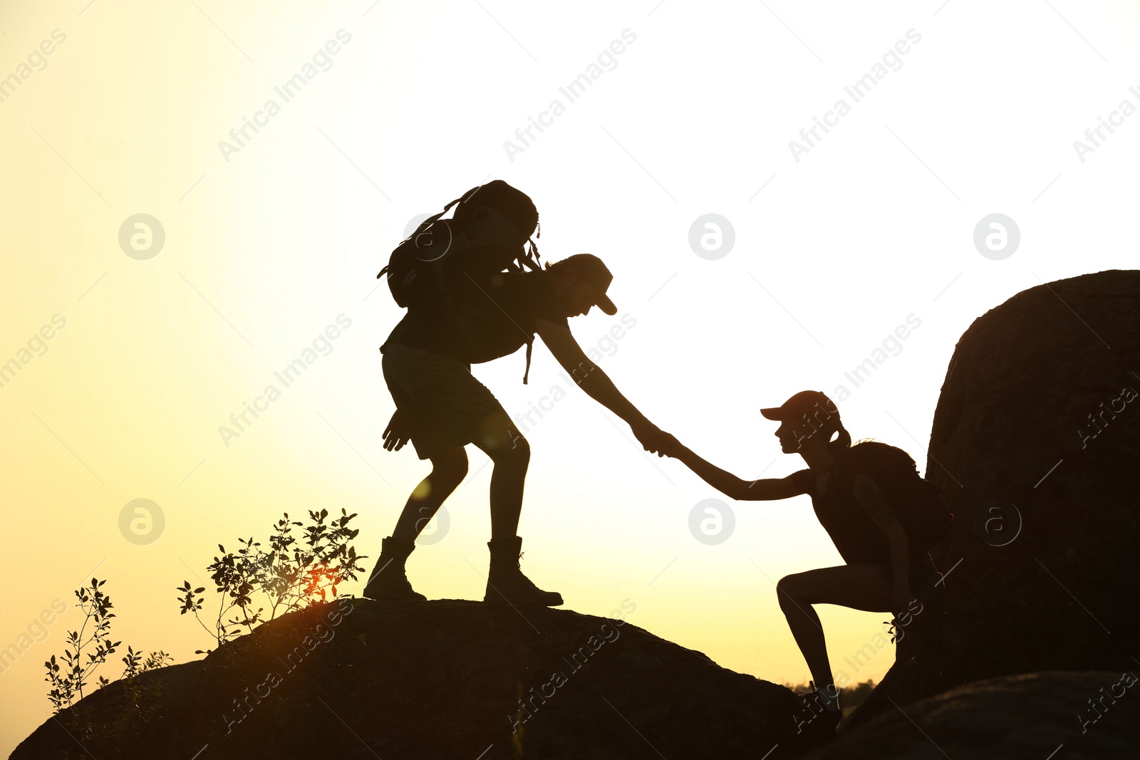 Photo of Silhouettes of man and woman helping each other to climb on hill against sunset