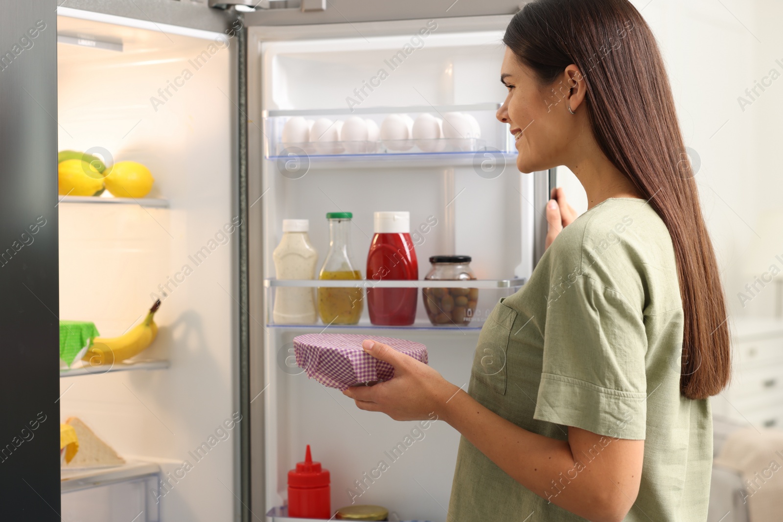 Photo of Happy woman holding bowl covered with beeswax food wrap near refrigerator in kitchen