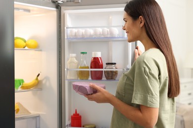 Happy woman holding bowl covered with beeswax food wrap near refrigerator in kitchen