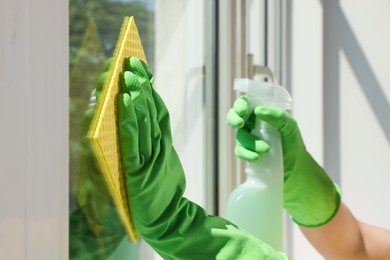 Photo of Woman cleaning window glass with sponge cloth and spray indoors, closeup