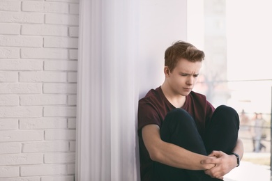 Upset teenage boy sitting alone near window indoors