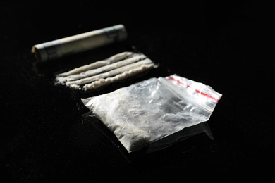 Photo of Drug addiction. Plastic bag with cocaine and rolled dollar banknote on black table, closeup