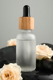 Photo of Bottle of face serum with beautiful roses on spa stones against grey background, closeup