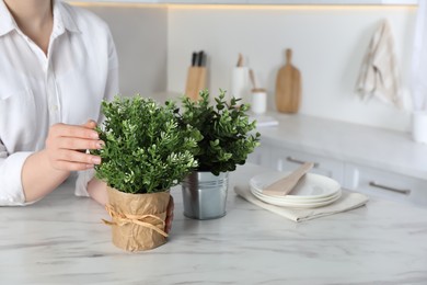 Photo of Woman near white marble table with different artificial potted herbs in kitchen, closeup. Space for text