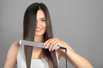 Photo of Young woman using hair iron on grey background, space for text