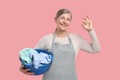 Photo of Happy housewife with basin full of laundry showing ok gesture on pink background