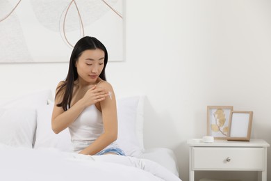 Beautiful young Asian woman applying body cream on shoulder in bedroom, space for text