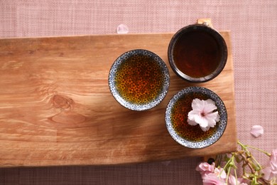 Photo of Wooden tray with cups and sakura flowers on pink fabric, flat lay. Traditional tea ceremony