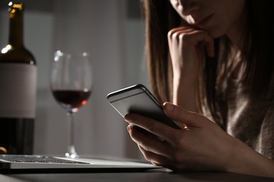 Photo of Woman using smartphone at table with laptop near glass of wine indoors, closeup. Loneliness concept