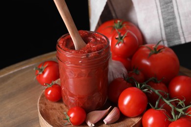 Photo of Jar of tasty tomato paste with spoon and ingredients on wooden table