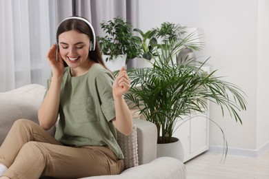 Beautiful young woman with headphones listening music on sofa in room with green houseplants
