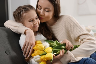 Photo of Happy woman kissing her cute daughter with bouquet of tulips on sofa at home