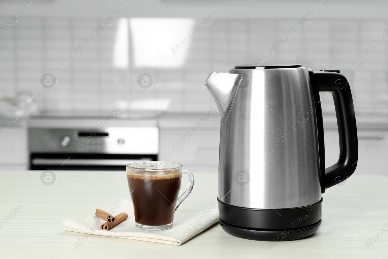 Photo of Modern electric kettle, cup of coffee and cinnamon sticks on white wooden table in kitchen