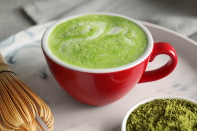 Photo of Cup of tasty matcha latte, green powder and bamboo whisk on tray, closeup