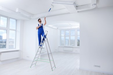 Photo of Handyman painting ceiling with roller on step ladder in room