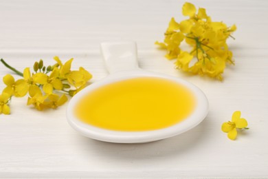 Photo of Rapeseed oil in gravy boat and beautiful yellow flowers on white wooden table, closeup