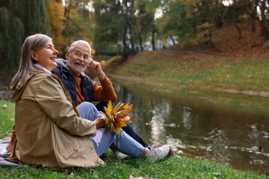Photo of Affectionate senior couple with dry leaves near river in autumn park, space for text
