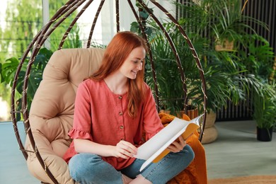 Photo of Happy young woman reading magazine in egg chair at indoor terrace