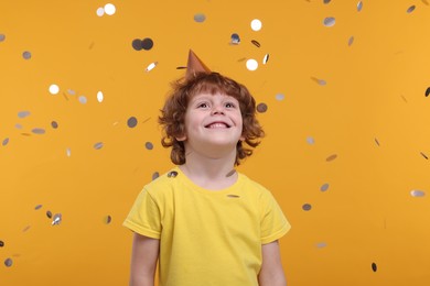 Photo of Happy little boy in party hat under falling confetti on orange background