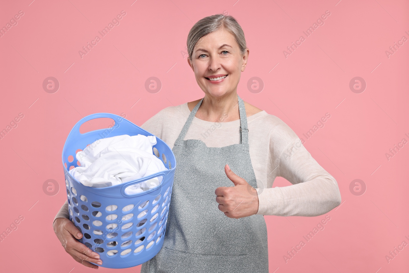 Photo of Happy housewife with basket full of laundry showing thumbs up on pink background