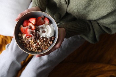 Woman holding bowl of tasty granola with chocolate chips, strawberries and yogurt indoors, top view