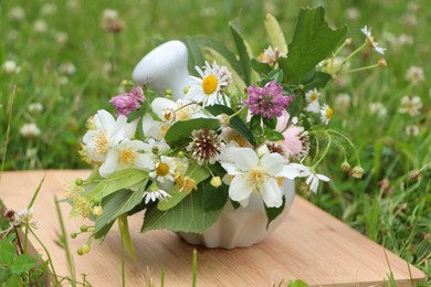 Ceramic mortar with pestle, different wildflowers and herbs on wooden board in meadow