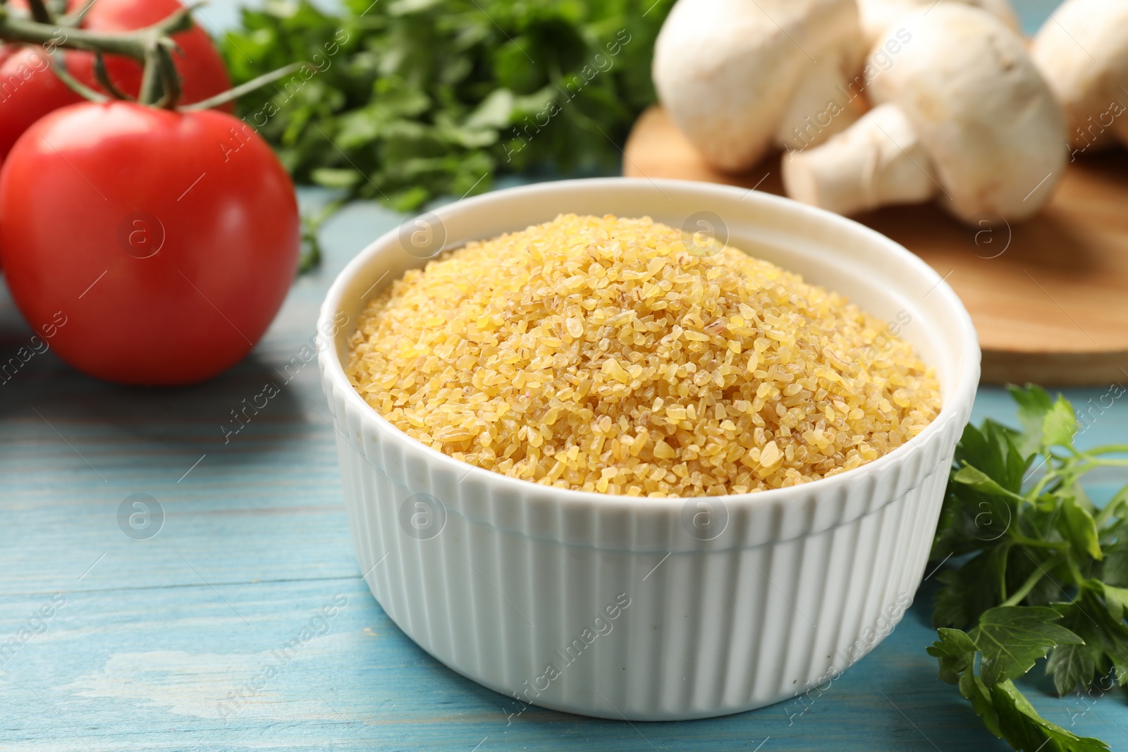 Photo of Raw bulgur in bowl, vegetables and parsley on light blue wooden table, closeup