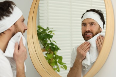 Washing face. Man with headband and towel near mirror in bathroom