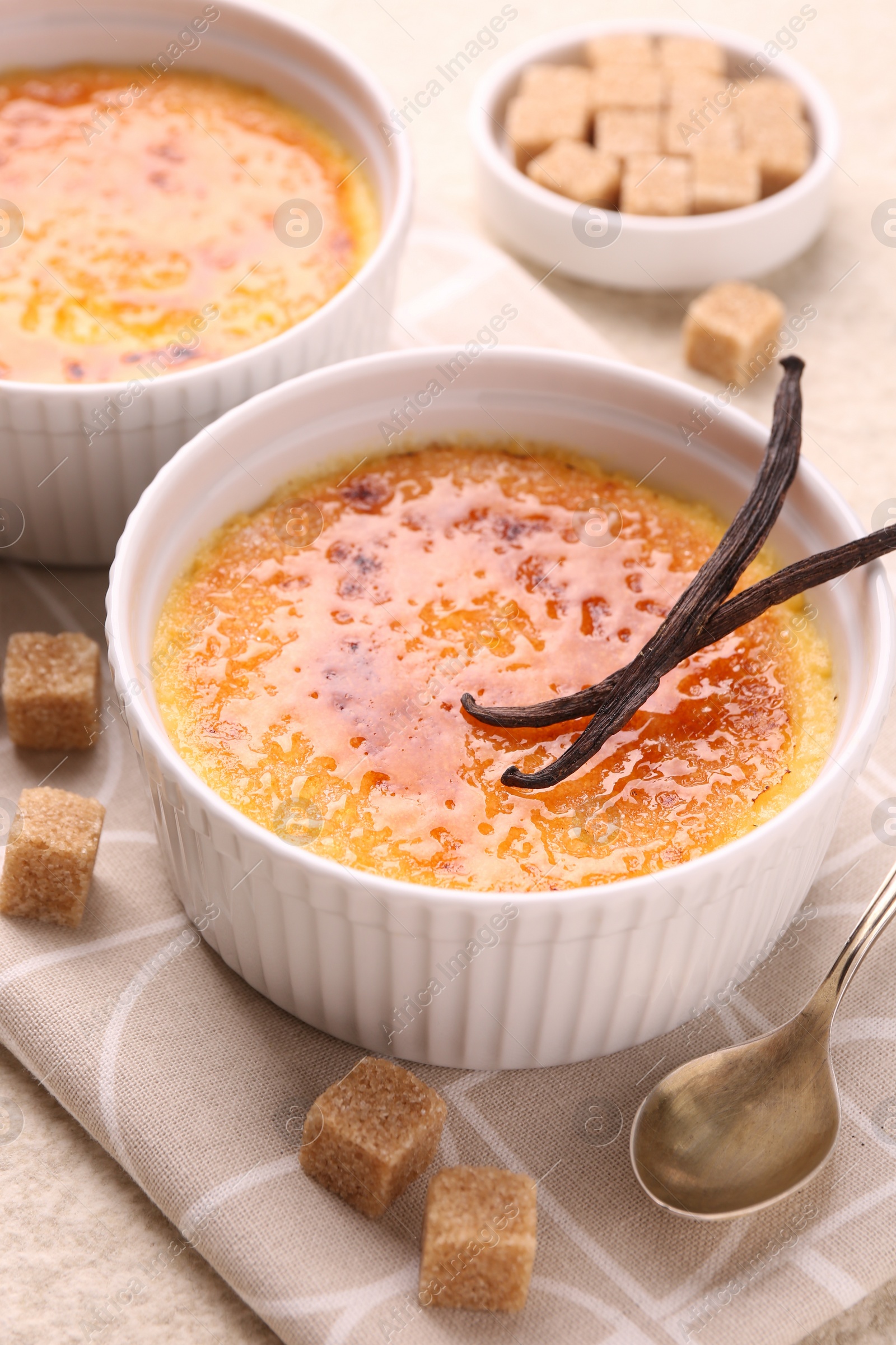Photo of Delicious creme brulee in bowl, vanilla pods, sugar cubes and spoon on table, closeup